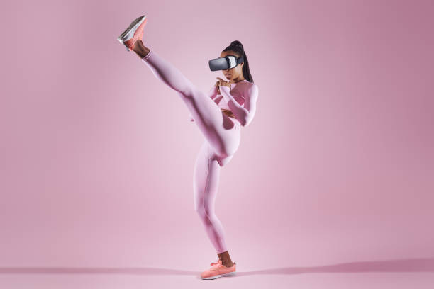 Woman in virtual reality headset practicing in kicking while standing against pink background Woman in virtual reality headset practicing in kicking while standing against pink background women boxing sport exercising stock pictures, royalty-free photos & images