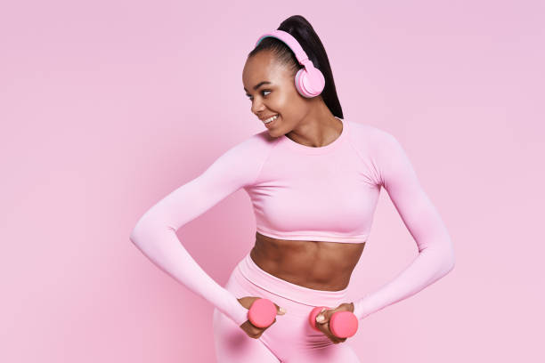 Attractive African woman in headphones exercising with dumbbells against pink background stock photo