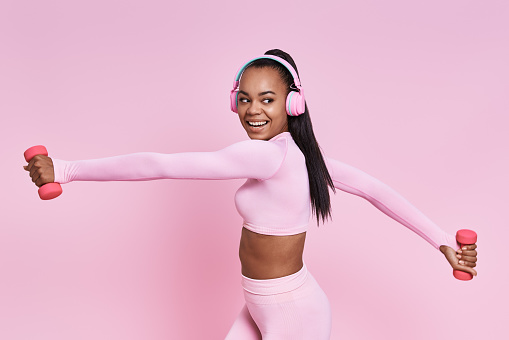 Cheerful African woman in headphones exercising with dumbbells against pink background