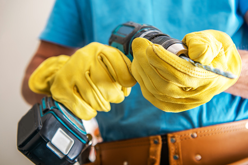 Closeup of Electrical Drill in Hands of Professional Construction Worker Wearing Yellow Protection Work Gloves. Professional Power Tools Theme.