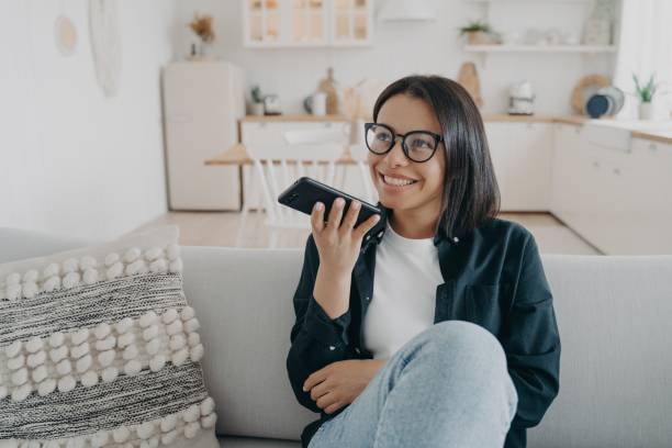 la femmina tiene lo smartphone, parla usando il vivavoce, registra il messaggio vocale, seduta sul divano di casa - conference phone immagine foto e immagini stock