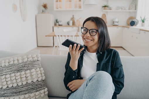 Smiling woman wearing glasses hold smartphone, chatting, sitting on couch at home. Female use virtual assistant on phone, talks with friend using speaker phone, records voice message in social network