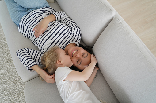 Kid kissing happy mother. Young mom having rest with her little kid on couch. European family is playing and relaxing together. Woman and daughter are enjoying leisure together. Togetherness and love.