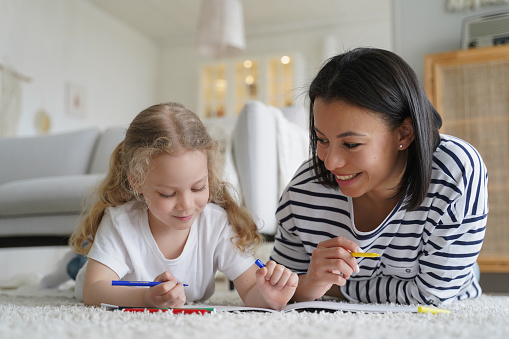 Smiling young mom and little foster daughter drawing painting together, lying on warm floor in living room. Mother or babysitter helps kid girl draw with colored pencils. Happy motherhood, adoption