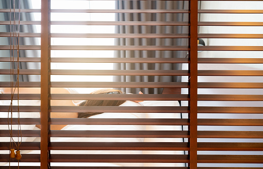 Wooden Venetian blinds. Blackout curtain between bathroom and bedroom in hotel with back view of woman sleep under white blanket on bed in the morning. Jalousie or window blinds. Horizontal blinds.