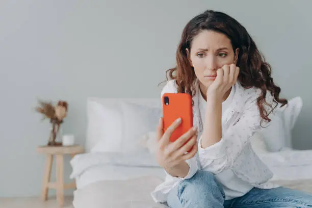 Photo of Stressed caucasian woman is doom scrolling. Girl reading morning news on smartphone in her bedroom.