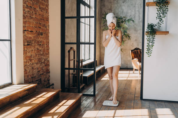 тревожная стройная кавказская женщина, завернутая в полотенце, делает шаг на весы, чтобы измерить свой вес. - sauna health spa healthy lifestyle wellbeing стоковые фото и изображения