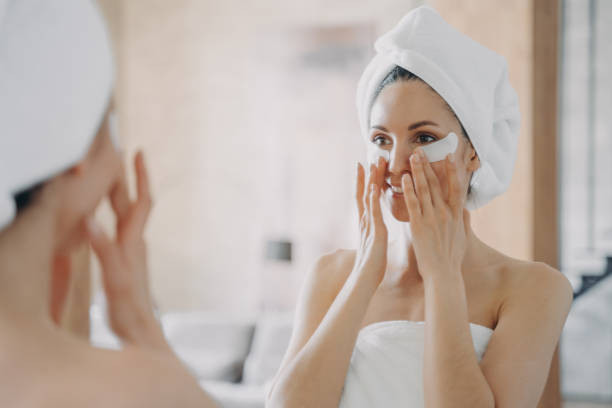 Girl applies eye patches and looking into mirror. Attractive woman wrapped in towel after bathing. Girl applies eye patches and looking into mirror. Attractive european woman wrapped in towel after bathing. Young hispanic lady takes shower at home. Relaxation at spa resort. Beauty routine at home. one eyed stock pictures, royalty-free photos & images