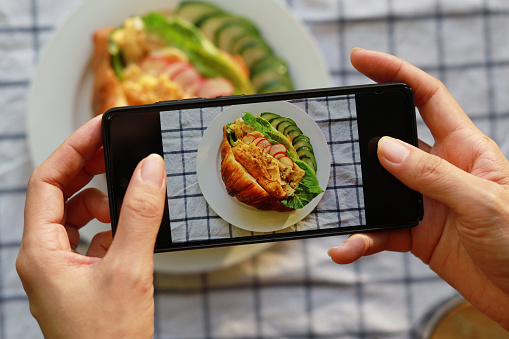 Woman 's hands taking a picture with smartphone of a plate of croissant sandwich served on a white plate