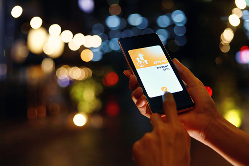 Cropped image of a woman's hand holding smartphone, sending money through digital wallet, bokeh illuminated light background. Smart banking with technology.