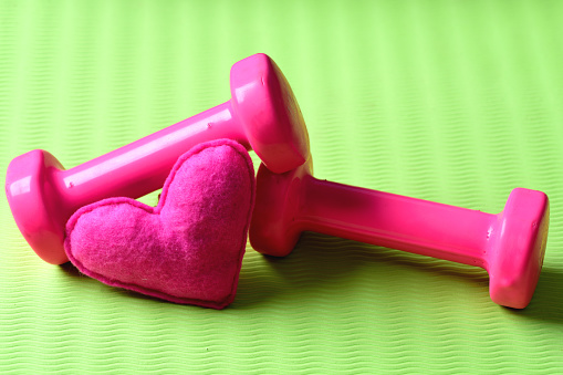 Love of sports and training. Heart decoration near plastic barbells on yoga mat. Dumbbells in pink color next to soft toy heart on green texture background, defocused. Female style and workout concept