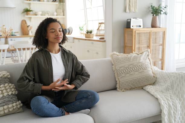 la ragazza biracial ama la meditazione, pratica lo yoga, seduta sul divano in posa di loto a casa - pranayama foto e immagini stock