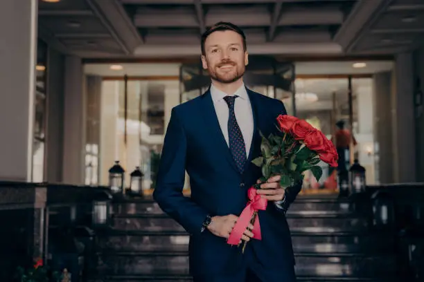 Happy smiling dapper dressed handsome businessman wearing blue suit, holding bouquet of fresh red roses with ribbon, waiting for girlfriend with smile, standing alone in front of hotel lobby entrance
