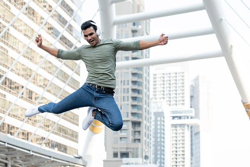 Full length of carefree Indian young man with arms outstretched jumping in mid-air while enjoying music over headphones against modern buildings in city