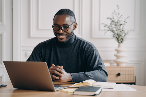 Positive African American male private consultant having session online listens to client attentively while looking at computer screen with smile, working remotely from home office. Freelance concept