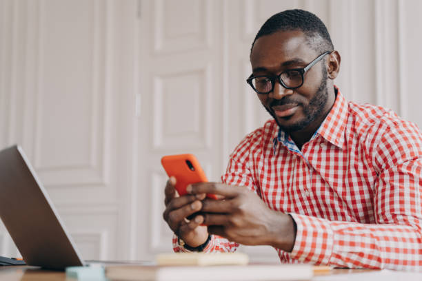 istock Young handsome african male executive in glasses sitting at office table and using mobile phone 1413449578