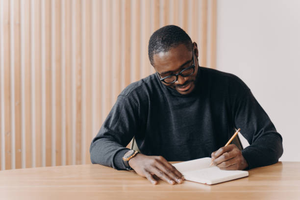 istock Focused young african man in glasses holding pen, taking notes in agenda while sitting at desk 1413449613