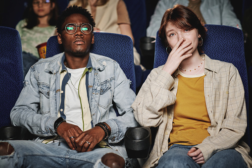 Young Black man and his Caucasian female friend watching sad drama movie in cinema, young woman crying