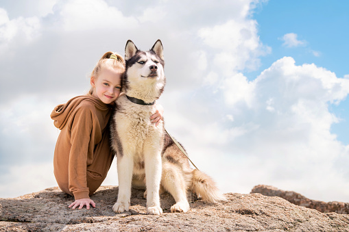 Siberian husky and his beautiful blue-eyed mistress are sitting on a rock against sky. The concept of love for animals and nature.