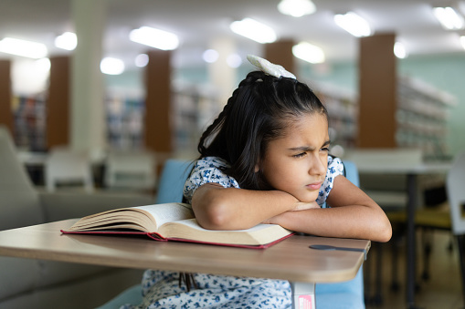 A little latin girl sitting at a desk with a book and frowning away.