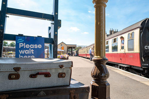 Shallow focus of bygone passenger luggage seen on a trolley prior to being loaded on the distant railway cars. The platform shows passengers boarding the train.