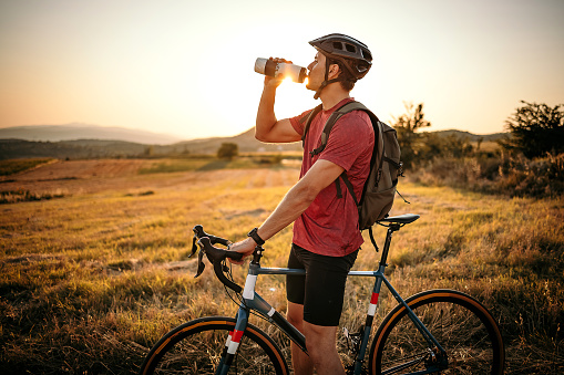 Young male athlete road cycling on a country road, taking a rest for refreshment, drinking water