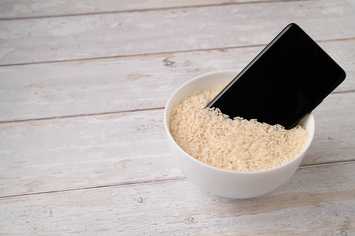 A mobile phone placed to dry in a bowl full of rice after the phone fell into water. Light wooden table background.