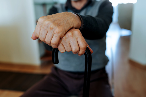A close-up of a senior Caucasian man sitting at home and holding a walking stick in his hands.