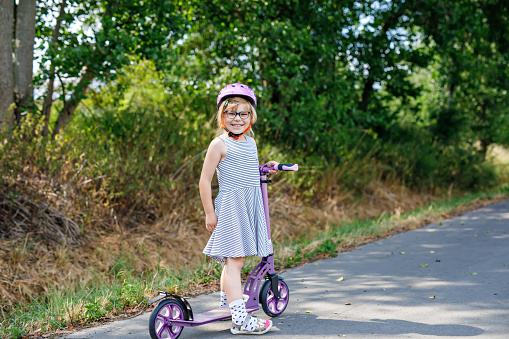 Portrait of active little preschool girl with glasses and helmet riding scooter on road in park outdoors on summer day. Seasonal child activity sport. Healthy childhood lifestyle.