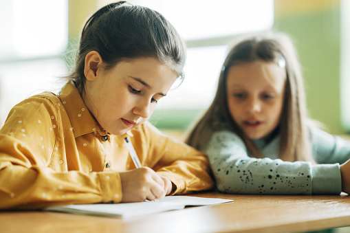 Cute schoolgirl writing in her workbook and her classmate looking into her book while sitting at desk in elementary school classroom. Schoolgirls sitting at desk and studying in classroom.