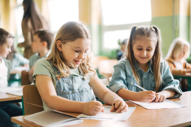 istock Smiling elementary schoolgirls writing exam at desk in classroom 1413476481