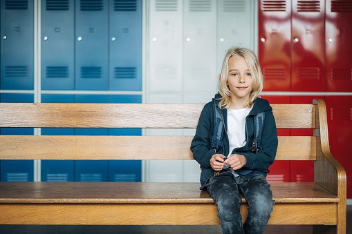 Portrait of elementary schoolboy sitting on bench in school corridor. Cute boy sitting alone on a wooden bench and looking at camera.
