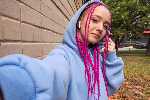 Portrait of a smiling caucasian teenage hipster girl with pink braids taking photo of herself, selfie,against a pink brick wall.Summer and technology concept.Generation Z style.Copy space.