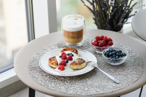 Cottage cheese pancakes for breakfast with blueberries and raspberries and sour cream. Cup of latte with delicious breakfast. Table with breakfast by the window on the background of the city.