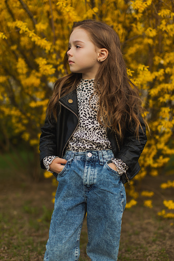 Little cute girl with long blonde curly hair in nature. Yellow flowering bush background. Beauty makeup with glitter. Childhood. Stylish child. Princess. Trendy clothes. Smell flowers. Allergy
