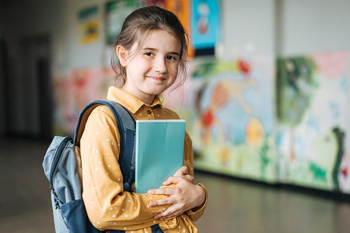 Portrait of happy young student with backpack and book standing in school corridor. Elementary schoolgirl looking at camera and smiling.