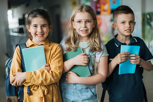 Portrait of happy elementary school children standing together in school corridor. Schoolgirls and schoolboy holding books.