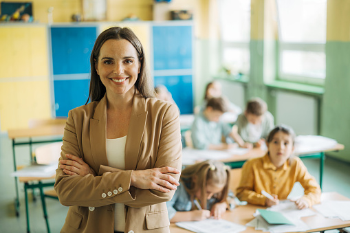 Portrait of young female teacher looking at camera and smiling while standing in front of her class in elementary school. Smiling woman teacher in elementary school classroom.