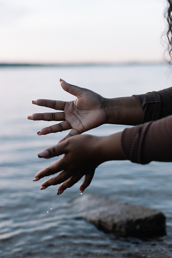 Cropped vertical of dark skin woman gently touching and splashing water drops in sea or river by hand. Blue sky horizon on background. Multicultural female on the beach. freshness and calm. Copy space