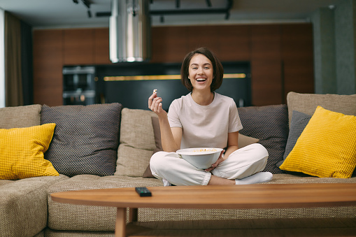 Laughing girl watching stand up comedy, comedian show, sitting with popcorn before television on sofa, rests at home on weekend. High quality photo