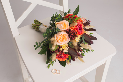 Wedding rings lie on white chair against background of bouquet. Wedding ceremony. Studio photography. Copy space