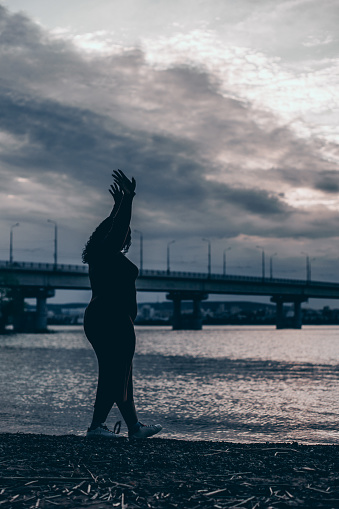 Vertical silhouette of curly serene overweight plump woman strolling river, sea beach with raised arms. Summer weekends against cloud sky horizon, bridge. Glare waves and clear water in summer evening
