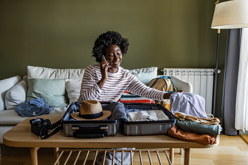 Portrait of young woman sitting on sofa bed and talking on mobile phone. She is packing her suitcase to go on vacation.