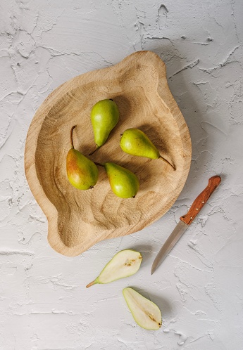 Istanbul, Turkey-August 6, 2022: Four green small pears on a large rustic wooden plate on a white plastered floor. Next to the plate is a pear cut in the middle and a cutting knife with a wooden handle. Shot with Canon EOS R5.