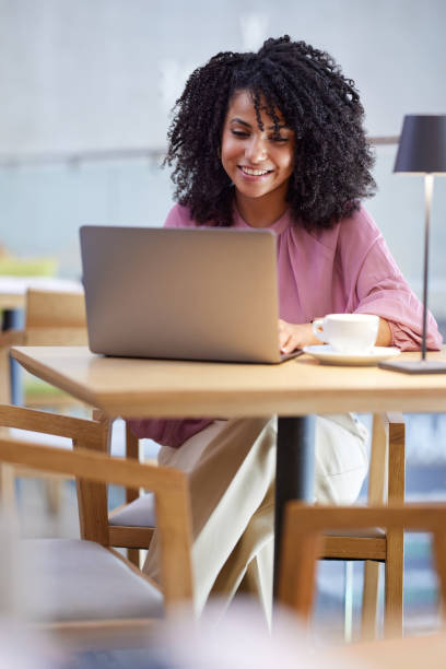 una feliz freelancer se sienta en una cafetería, escribiendo en un teclado mientras sonríe a la computadora portátil. - typebar fotografías e imágenes de stock