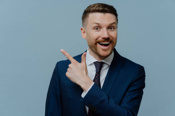 istock Happy successful male entrepreneur with bristle dressed in formal clothes points at upper right corner demonstrated place for your advertisement gives advice how to start own business. Office style 1413493824