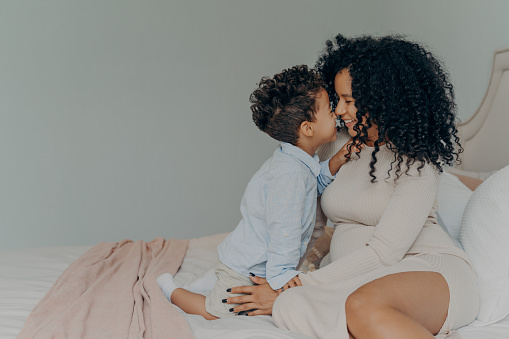 Tender moment of young family, afro american pregnant mother and son sitting on bed,touching each other with noses, white wall background in bedroom interior. Happy parenting and motherhood concept