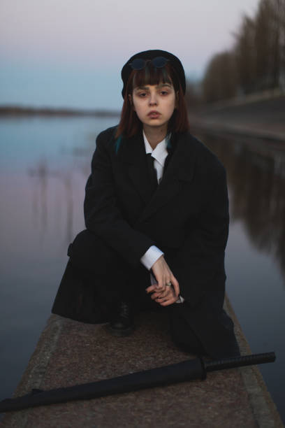 fille rousse en vêtements noirs sur fond de rivière à l’extérieur. style gothique. modèle asiatique en manteau sombre avec parapluie sur la nature. surface de l’eau. tenue élégante. style moderne pour les jeunes. sous-culture. art - gothic style photos et images de collection