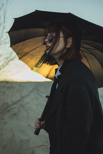 Redhead girl in black clothes on sunset background outdoors. Gothic style. Asian fashion model in dark coat with umbrella on nature. Black sunglasses. Stylish outfit. Youth modern style. Subculture
