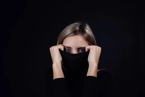 Anxious woman hiding face with black blouse collar with eyes looking at camera on black background. Victim of physical and psychological abuse. Gaslighting. Relative aggression.